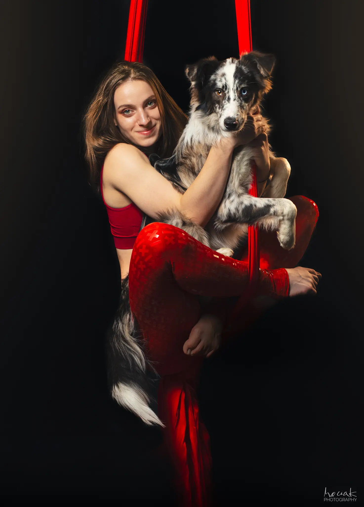 Zbliżenie na kobietę wykonującą akrobację na czerwonej szarfie, która trzyma w ramionach psa rasy border collie. Pies ma charakterystyczne, różnokolorowe oczy (jedno brązowe, drugie niebieskie). Oboje patrzą w stronę aparatu na ciemnym tle.