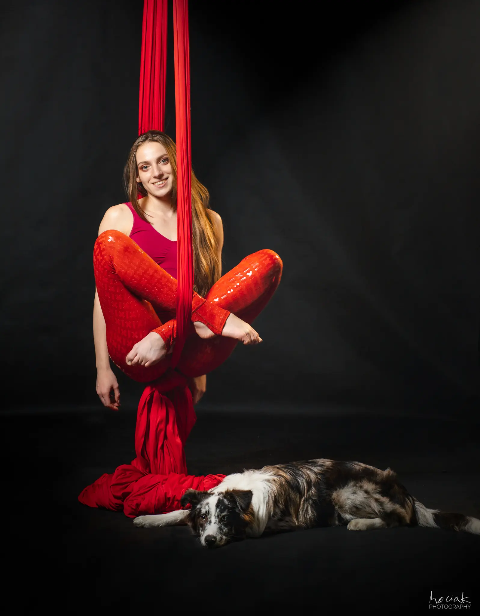 Kobieta o długich brązowych włosach siedzi w powietrzu na czerwonej szarfie akrobatycznej na czarnym tle. Ma na sobie czerwony strój sportowy, a jej nogi są zaplecione w materiale. Na podłodze pod nią leży spokojnie pies rasy border collie o umaszczeniu blue merle.