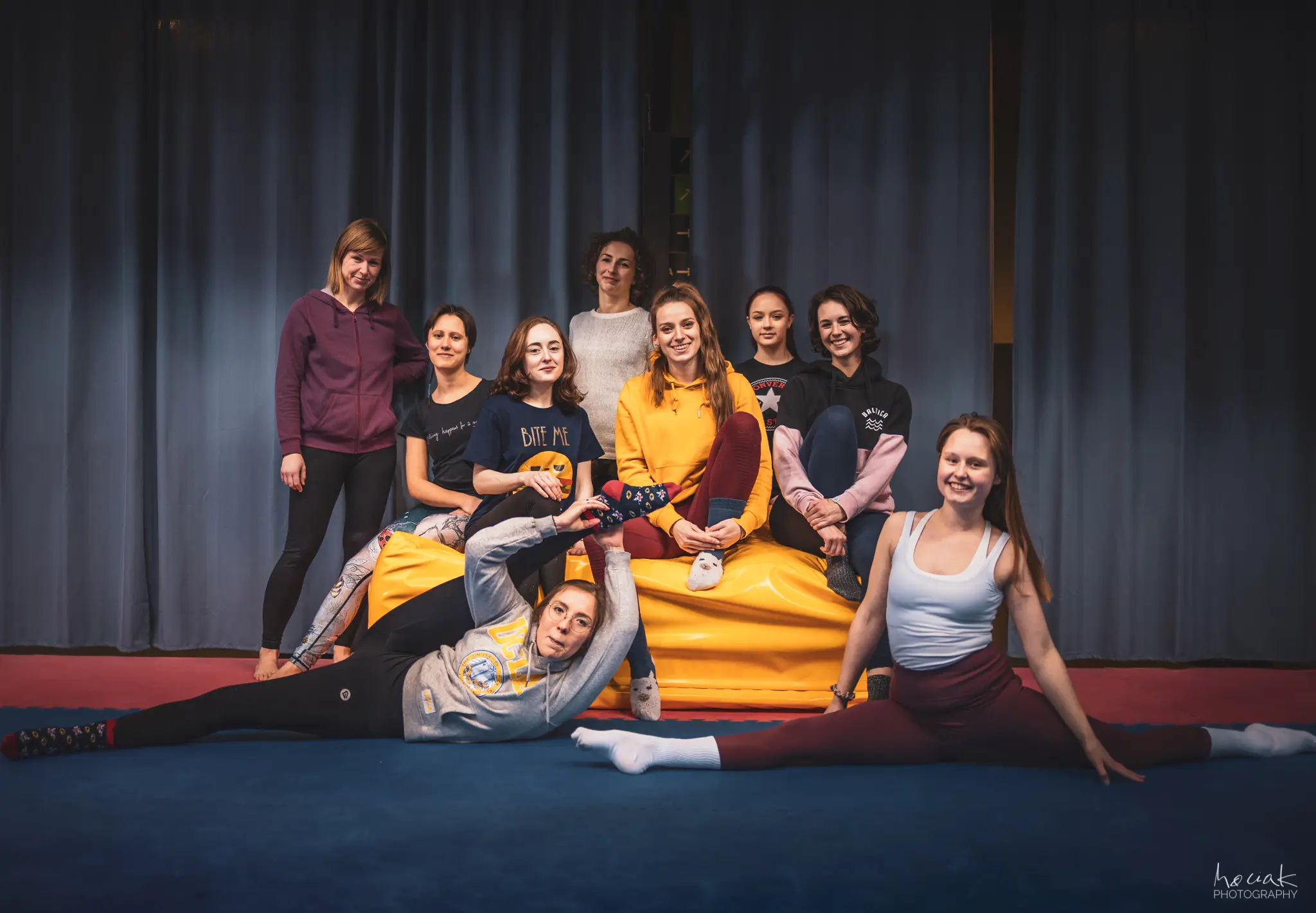 Grupa uśmiechniętych kobiet pozuje wspólnie na sali gimnastycznej po warsztatach akrobatycznych. Dwie uczestniczki na pierwszym planie prezentują szpagaty na niebieskiej macie, podczas gdy reszta grupy siedzi na żółtym materacu i stoi w tle na tle ciemnej zasłony.
