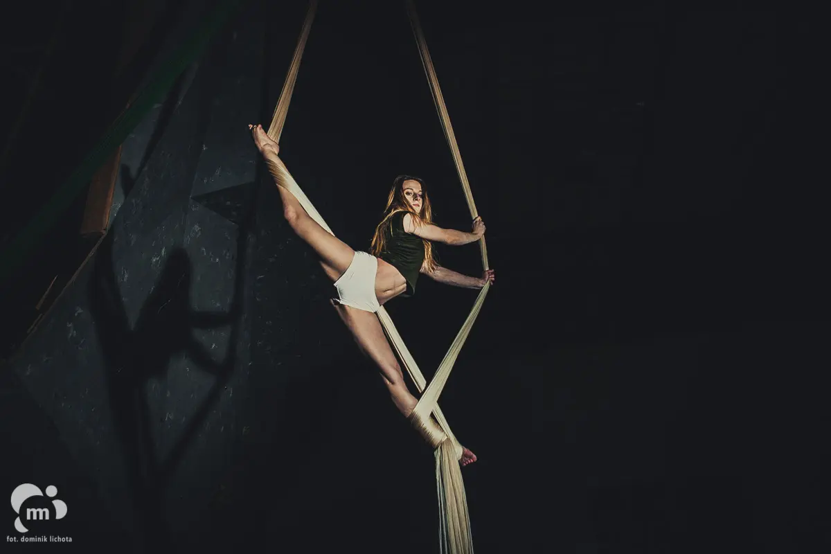 Kobieta wykonująca akrobacje na szarfach w powietrzu (aerial silks) na ciemnym tle.