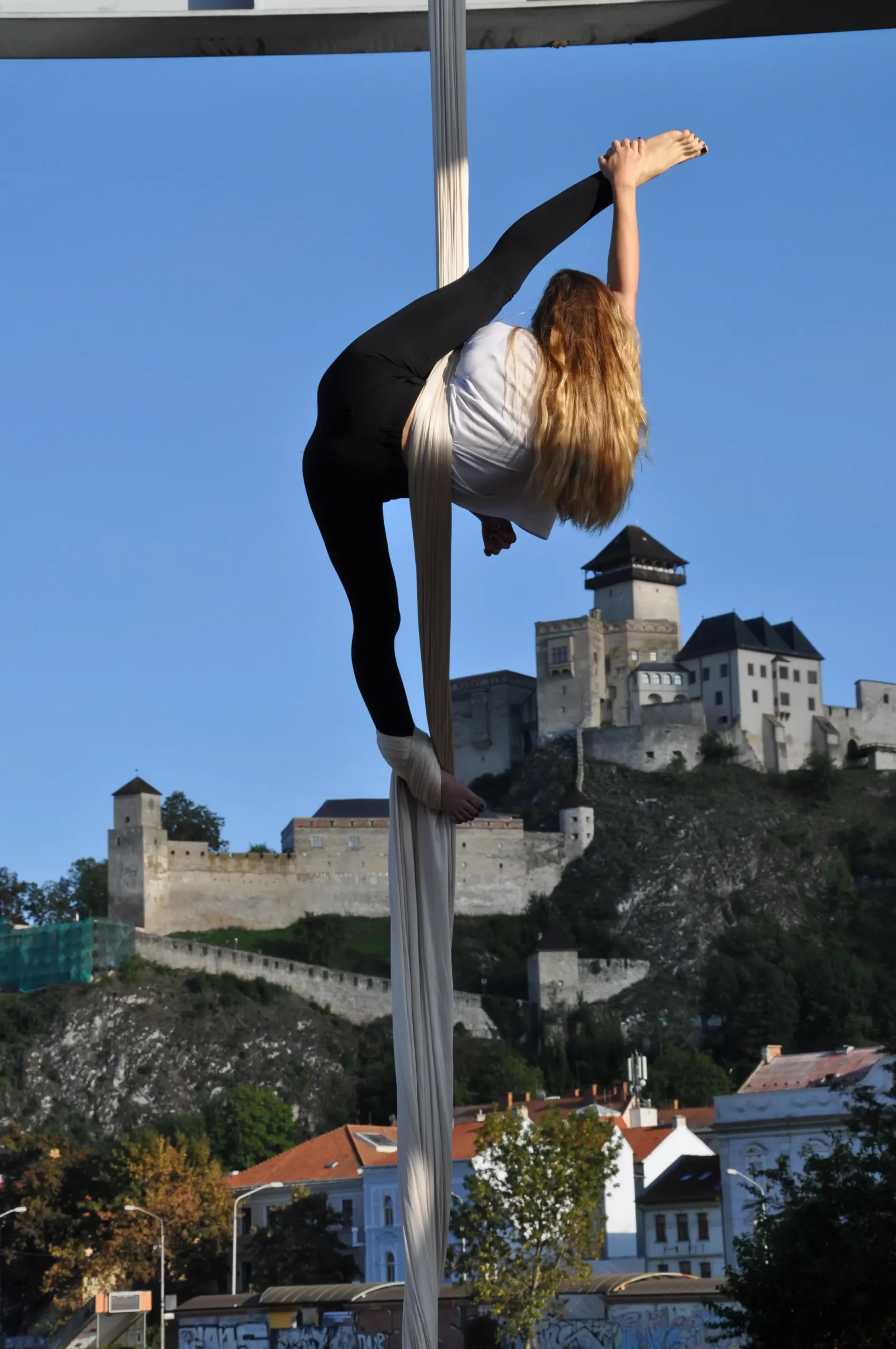Klaudia Bawer wykonująca zaawansowaną figurę na białej szarfie akrobatycznej na tle historycznego zamku i błękitnego nieba.