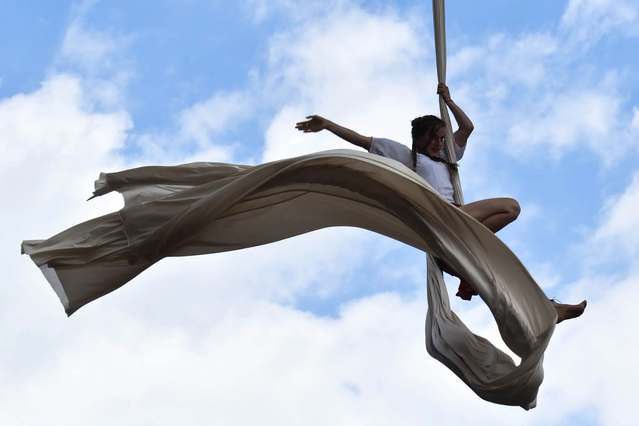 Klaudia Bawer w dynamicznej figurze na szarfach akrobatycznych na tle błękitnego nieba z chmurami. Długi, beżowy materiał malowniczo powiewa na wietrze.