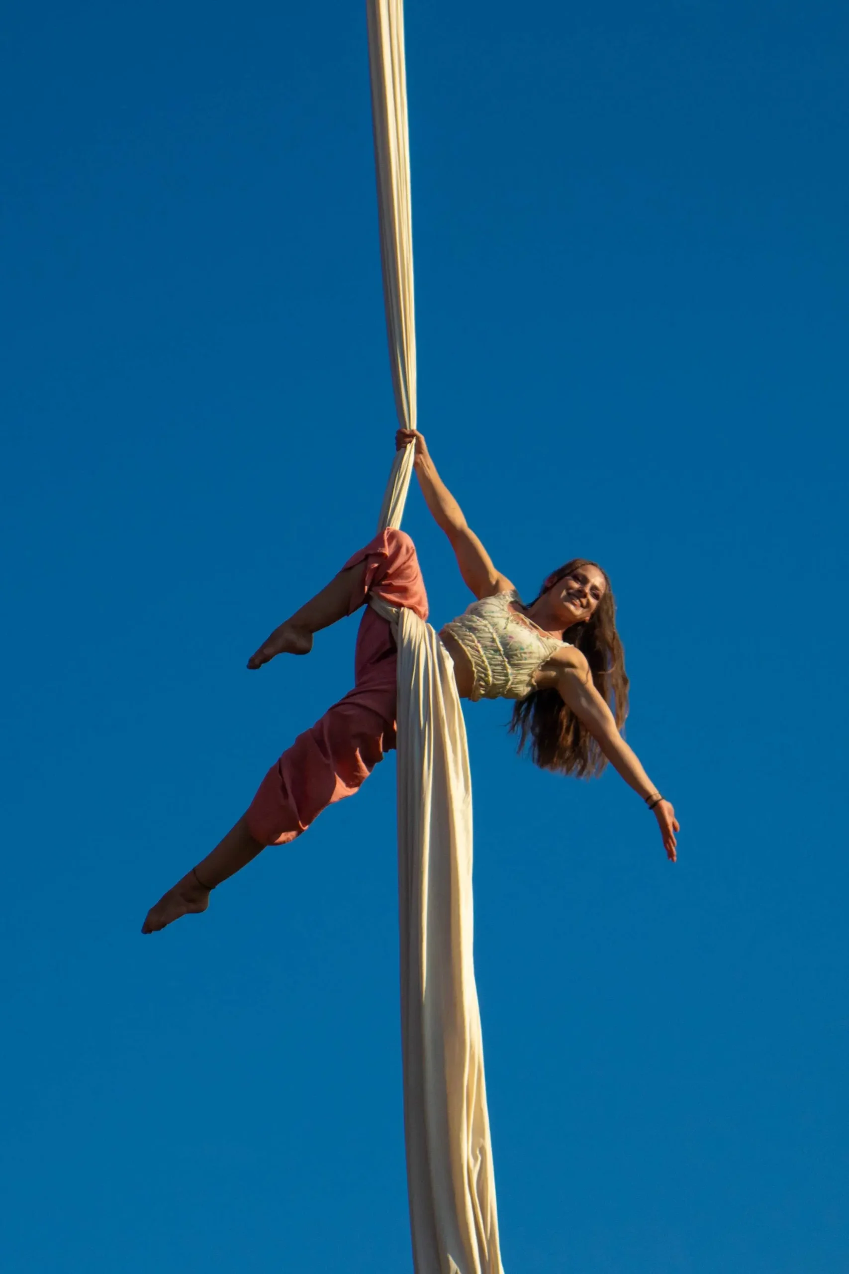 Uśmiechnięta akrobatka wykonująca figurę na białej szarfie podwieszonej na zewnątrz. Zdjęcie zrobione od dołu na tle intensywnie niebieskiego nieba.