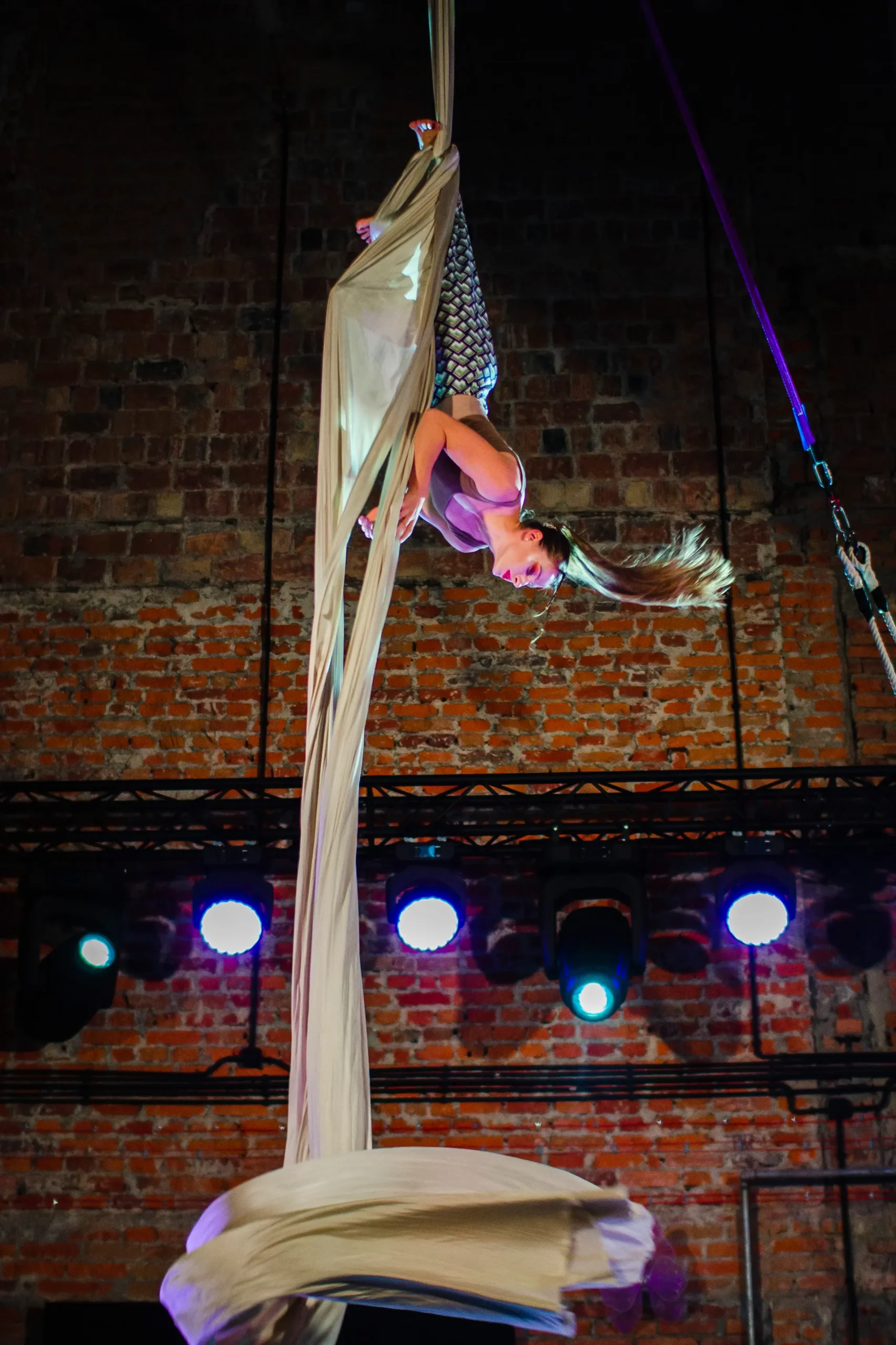 Klaudia Bawer wykonuje akrobację głową w dół na kremowej szarfie (aerial silks). W tle surowa ściana z cegieł oraz niebieskie reflektory sceniczne podkreślające dynamikę ruchu.